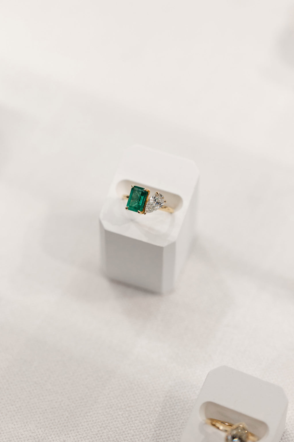 A gold ring with a rectangular green emerald-cut emerald and a pear cut diamond, displayed on a white jewelry stand against a soft white fabric background.