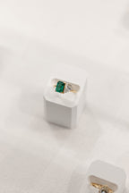 A gold ring with a rectangular green emerald-cut emerald and a pear cut diamond, displayed on a white jewelry stand against a soft white fabric background.