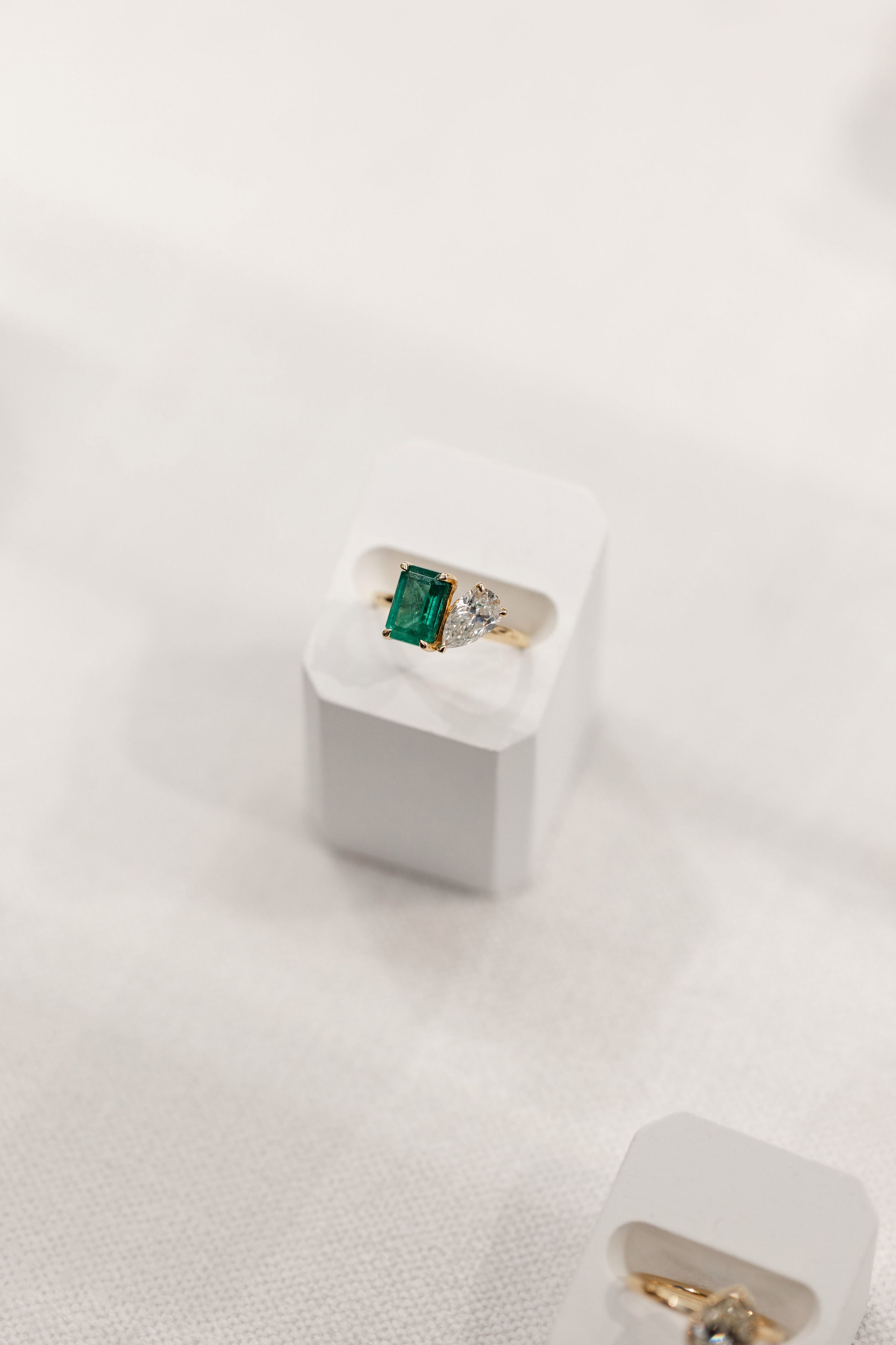 A gold ring with a rectangular green emerald-cut emerald and a pear cut diamond, displayed on a white jewelry stand against a soft white fabric background.