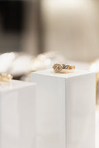 A gold signet ring with a marquise and small round diamonds, displayed on a white pedestal in a softly blurred jewelry setting.