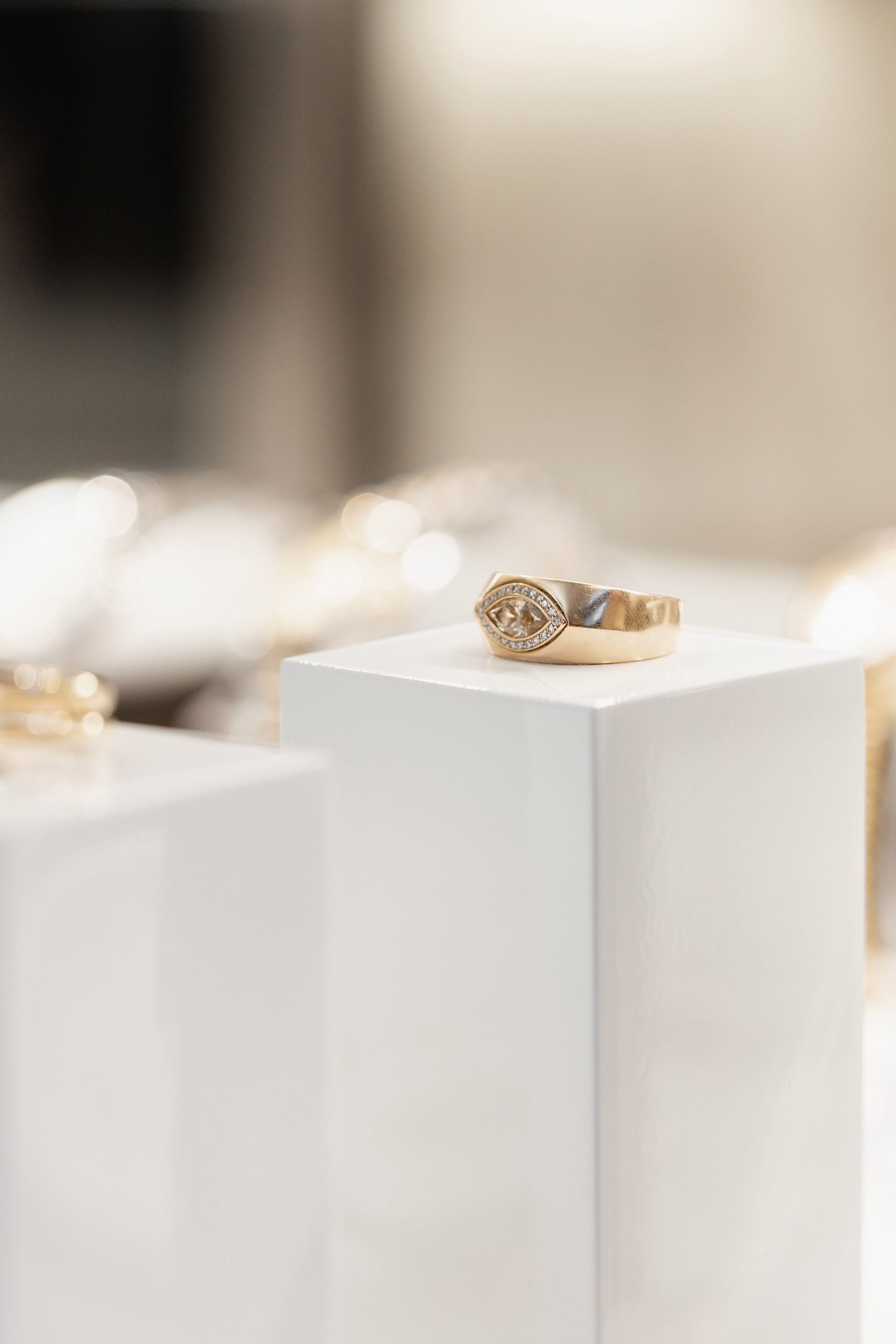 A gold signet ring with a marquise and small round diamonds, displayed on a white pedestal in a softly blurred jewelry setting.