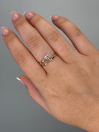 A close-up of a hand wearing a double-band ring set with a heart shaped diamond on one band and a slim yellow gold band on the other, against a plain gray background.