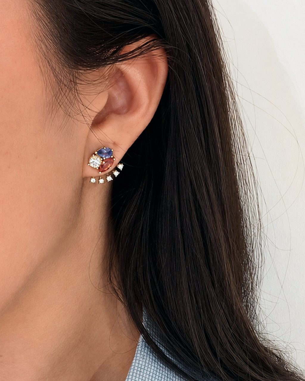 A close-up of a person's ear wearing a gold crescent-shaped ear cuff earring adorned with a blue pear shaped tourmaline, a pinkish-orange marquise garnet, and round diamond.