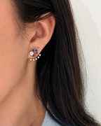 A close-up of a person's ear wearing a gold crescent-shaped ear cuff earring adorned with a blue pear shaped tourmaline, a pinkish-orange marquise garnet, and round diamond.