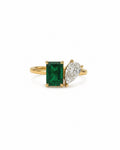 A yellow gold ring featuring a rectangular emerald-cut emerald and a pear-cut diamond on a white background.