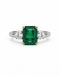 An emerald-cut green emerald ring with a white gold band and two round-cut diamonds on either side, set in gold-colored prongs, shown against a plain white background.